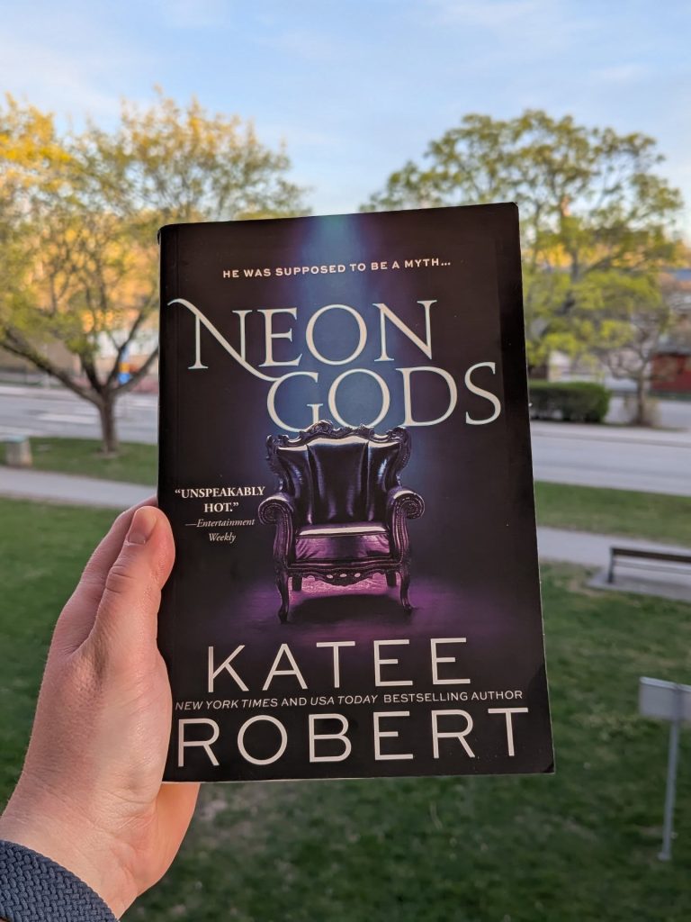 Bokomslaget till Neon Gods av Katee Robert, fotograferat mot en naturlig bakgrund med grönska.